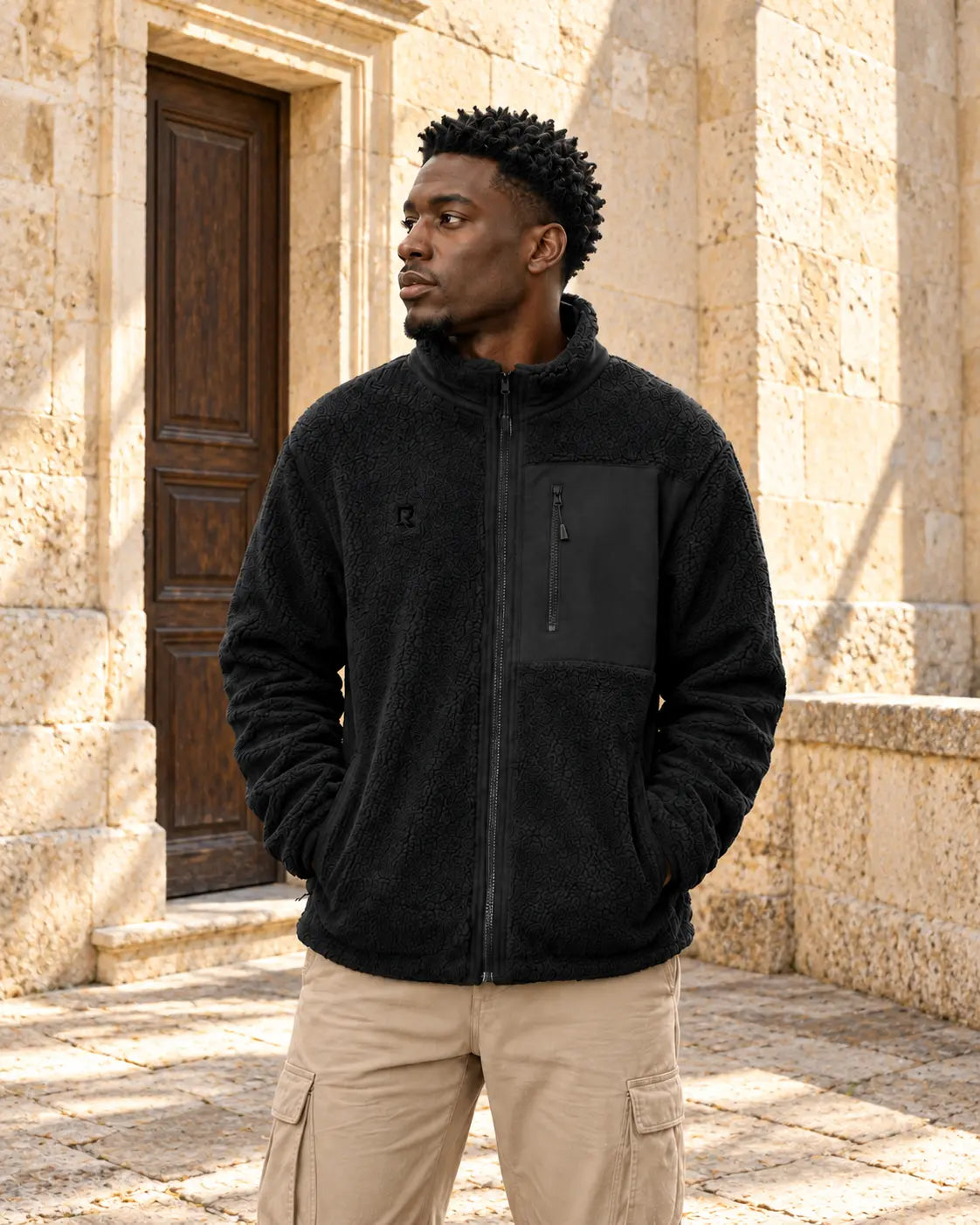 Veste Sherpa Le Refuge RIVHEX noire portée dans une cour en pierre claire, broderie ton sur ton